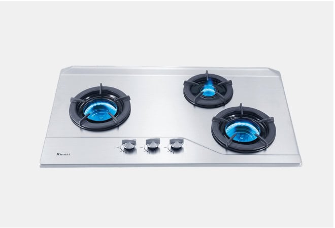 Stainless steel gas cooktop with three burners lit and blue flames, knobs centered toward the front, Rinnai branding visible on the left edge.