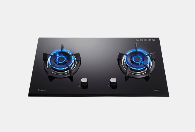 Two-burner black glass gas cooktop with blue flames lit and two central control knobs, modern kitchen appliance.