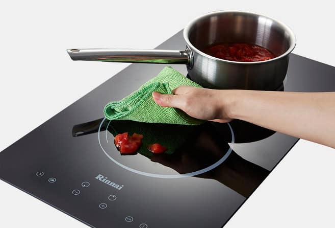 Hand wiping tomato residue off a black induction cooktop with a green cloth; a stainless pot sits on the burner.