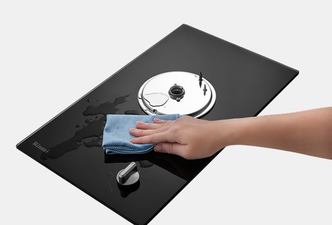 Person cleaning a black pool cleaning robot with a blue microfiber cloth, wiping around a round top panel.