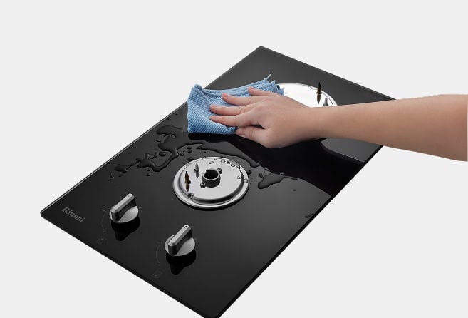 Hand wiping a black glass gas stove top with a blue cloth, water droplets on the surface and control knobs visible at the bottom left.