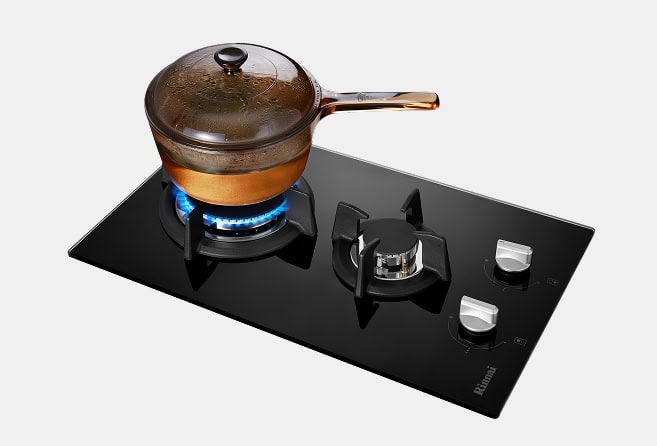 Copper pot with glass lid sitting on a lit gas burner of a black glass cooktop.