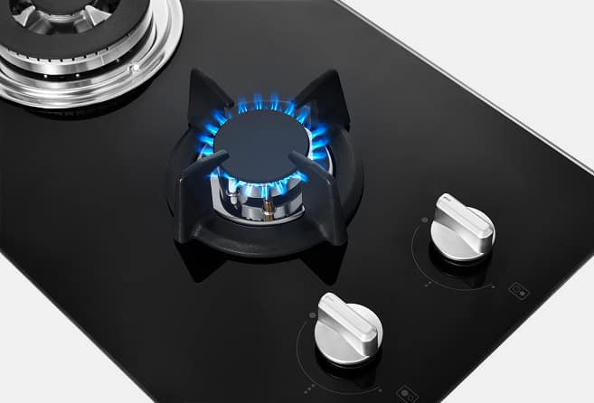 Gas stove with a lit burner and blue flame on a black glass cooktop, with two silver control knobs nearby in the foreground.