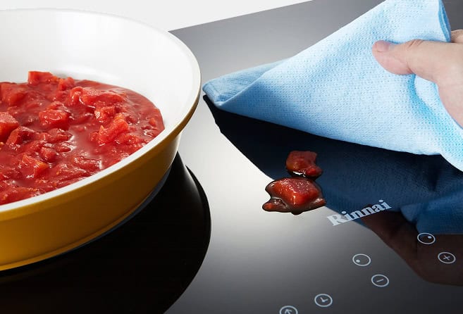 Yellow frying pan with tomato sauce simmering on a stove, beside a hand wiping a glossy induction cooktop with a blue cloth