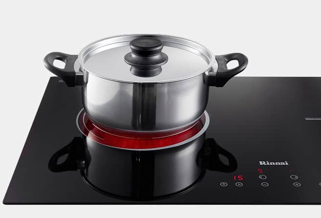 Stainless steel pot with glass lid on a glowing red induction burner on a black glass cooktop.