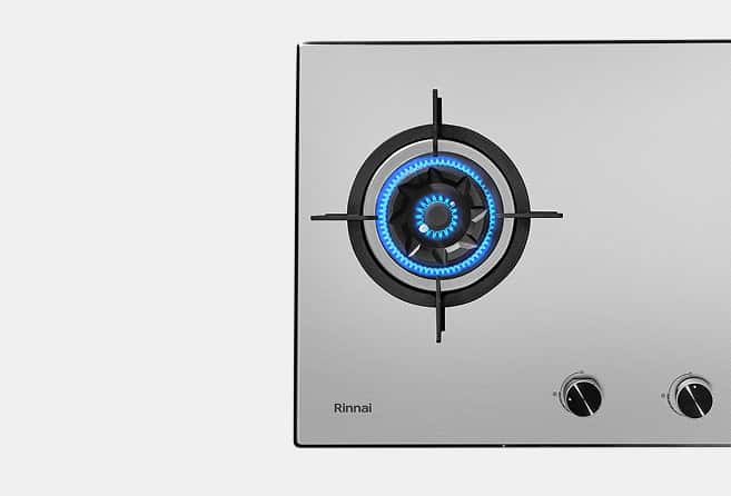 Stainless steel gas cooktop with a single lit burner showing a blue flame; Rinnai branding on the left and two control knobs on the right.