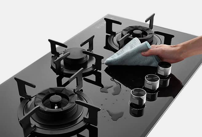 Person wiping a glass-top gas stove with a microfiber cloth, cleaning the surface around the burners