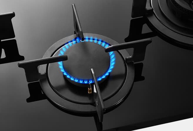 Lit gas stove burner with a bright blue flame around the burner cap and surrounding grate, on a dark cooktop surface.