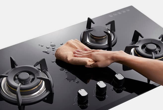 Person’s hand wiping a black glass gas stove top with a beige cloth, cleaning spills between burners.