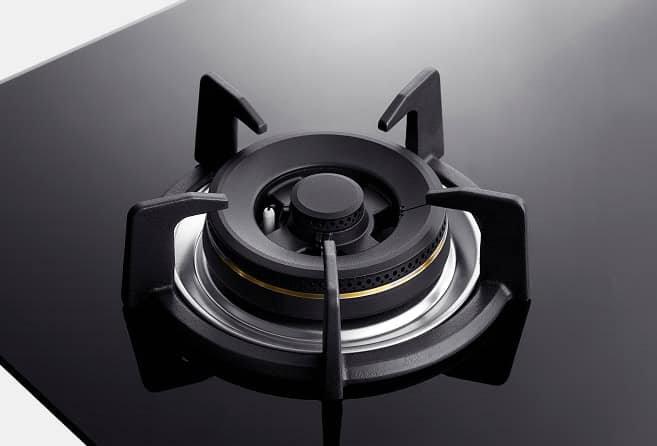 Top view of a gas stove burner on a black glossy surface, showing four metal pan supports and a central burner cap.