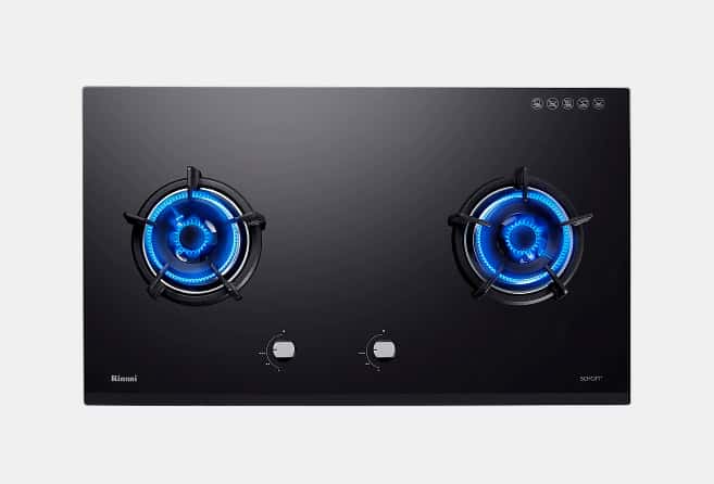 Black glass two-burner gas cooktop with blue flames lit on both burners and central control knobs.