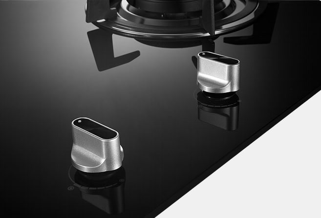 Close-up of two silver gas stove knobs on a glossy black cooktop, with a gas burner visible in the background.