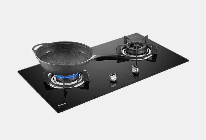 Black glass gas cooktop with two burners; a dark frying pan sits on the left burner over a blue flame.