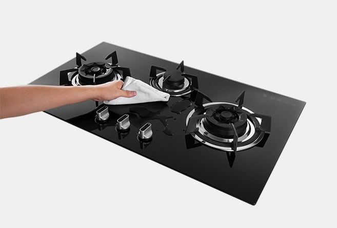 Hand wiping a three-burner black glass gas cooktop with a cloth for cleaning