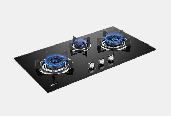 Black glass gas cooktop with three lit burners showing blue flames and stainless steel pan supports; control knobs in front.