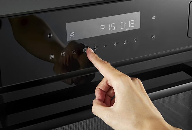 Person using a touchscreen control panel on a sleek kitchen appliance (likely an oven) in a modern kitchen surface nearby.