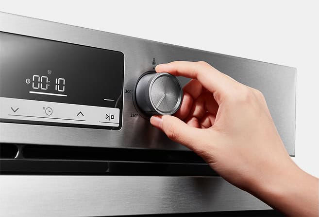 Hand turning the temperature knob on a stainless steel oven control panel; digital timer displays 00:10