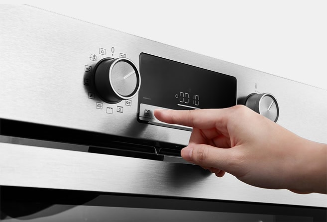 Hand pressing a button on a stainless steel oven control panel in a kitchen setting.