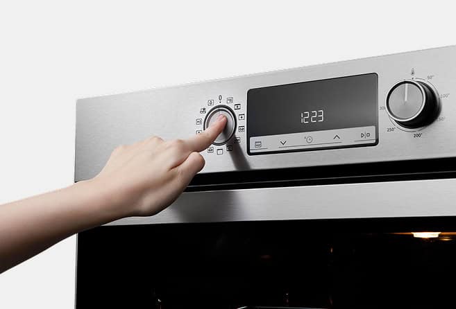 Person adjusting a stainless steel oven control panel by turning a dial next to a digital clock display showing 12:23, likely setting temperature or mode.