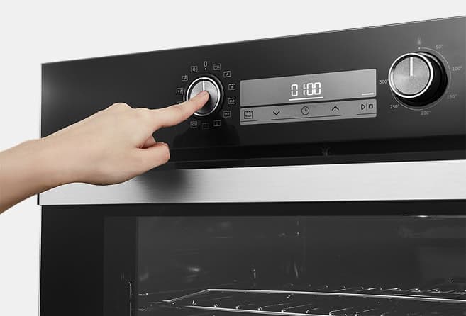 Close-up of a hand turning the left control knob on a modern oven with a digital timer display.
