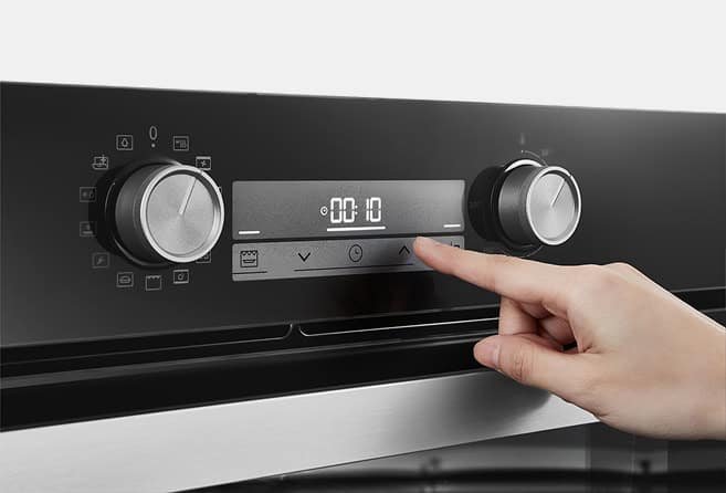 Close-up of a hand pressing the control panel on a modern oven, flanked by two large knobs.