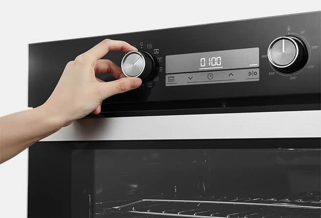 Close-up of a hand turning the oven's control knob on a black front panel with a digital display showing 0:00