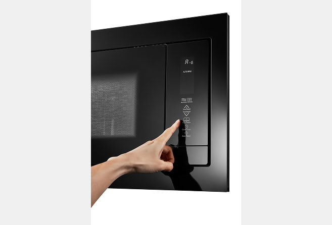 Hand pressing the control panel of a black microwave oven in a kitchen setting.