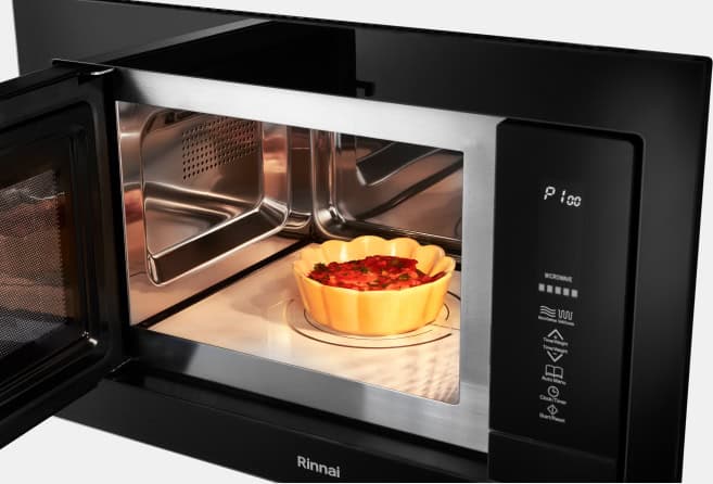 Black microwave oven with its door open, revealing a golden tart baking on a glass turntable inside. Popular interior lighting highlights the metal walls.",