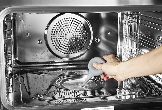 Person pours water from a squeeze bottle into a metal tray inside a stainless steel oven for cleaning/steam cleaning.