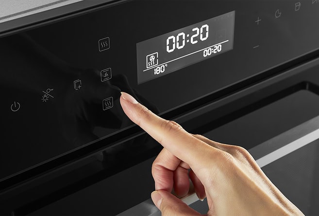 Hand pressing a low-profile button on a modern oven control panel, timer shows 00:20 and steam icon nearby (functional action).