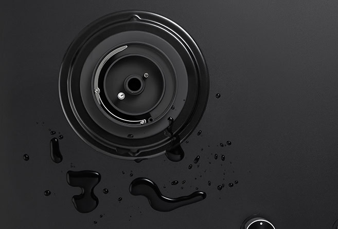 Close-up of a round metal lens mount with inner rings on a dark surface, with scattered black droplets nearby.